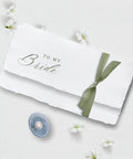 White handmade paper with 'To My Bride' text and a green ribbon, surrounded by small white flowers on a light background.