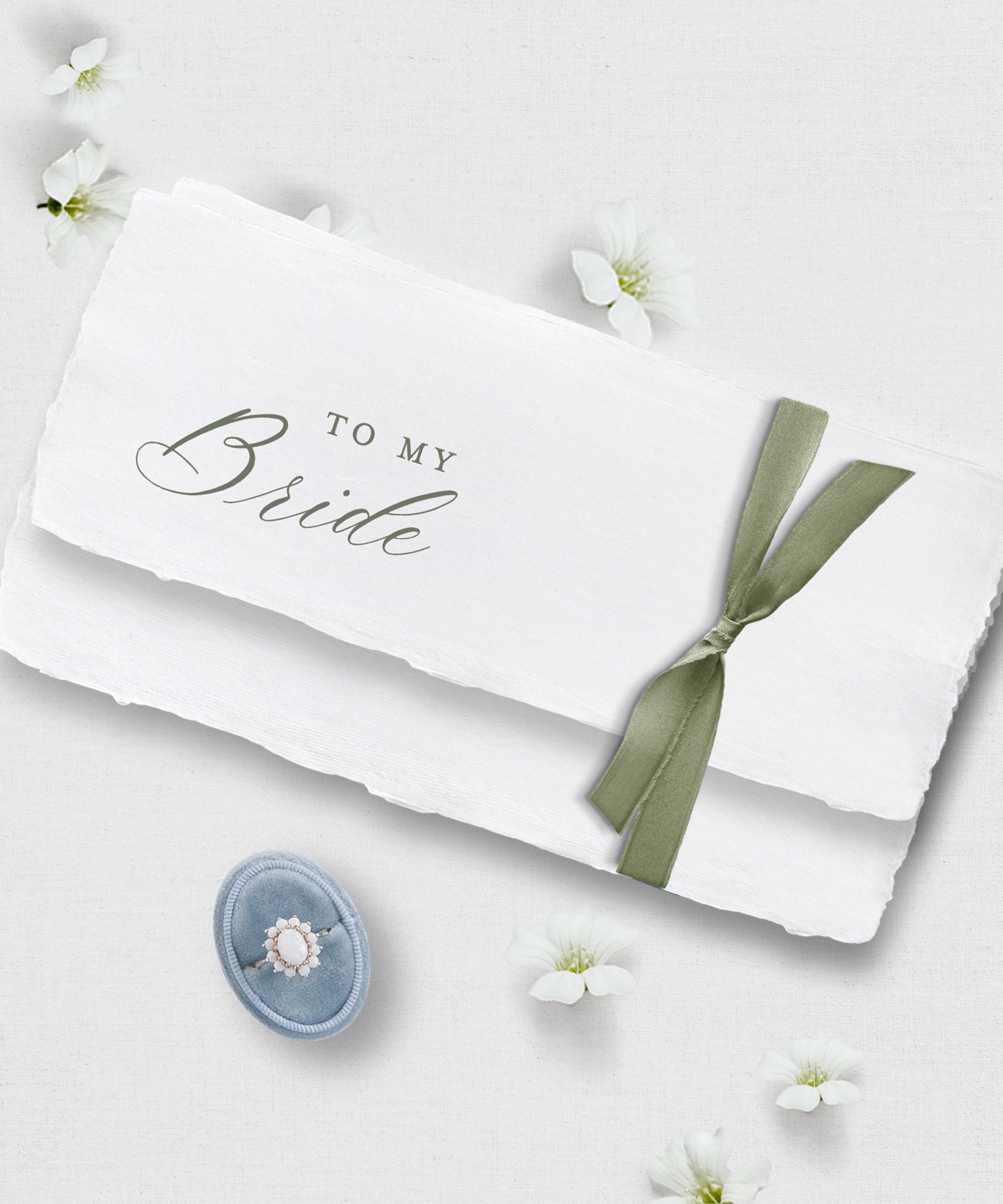 White handmade paper with 'To My Bride' text and a green ribbon, surrounded by small white flowers on a light background.