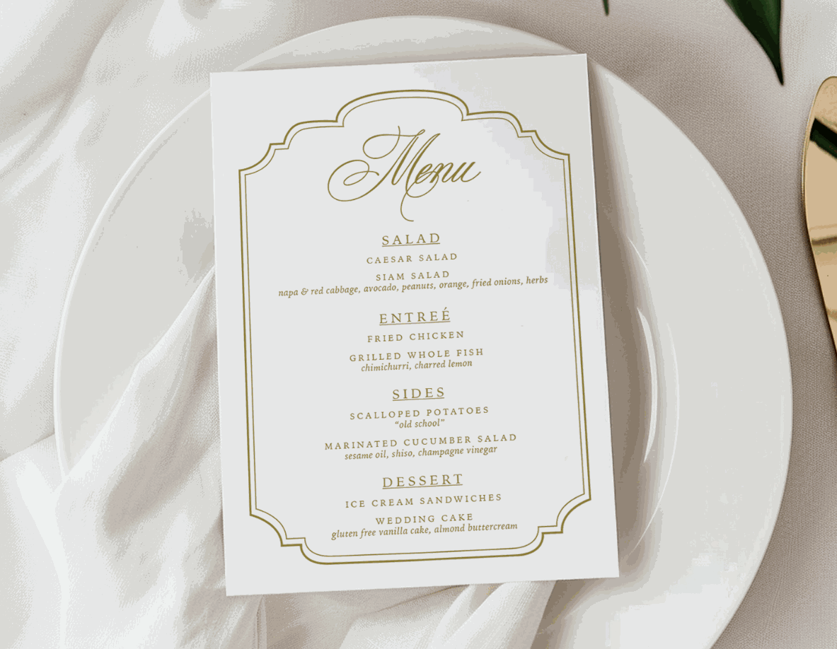Menu card on a white plate with gold accents, surrounded by flowers and cutlery on a white tablecloth.