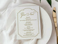White plate with a gold-rimmed menu card on a white tablecloth with flowers and cutlery.